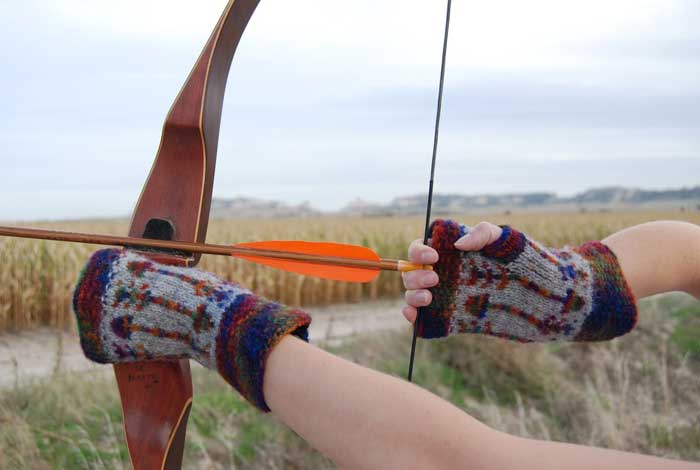 two hands holding bow and arrow poised for shooting and wearing colorwork fingerless mitts in an arrow motif