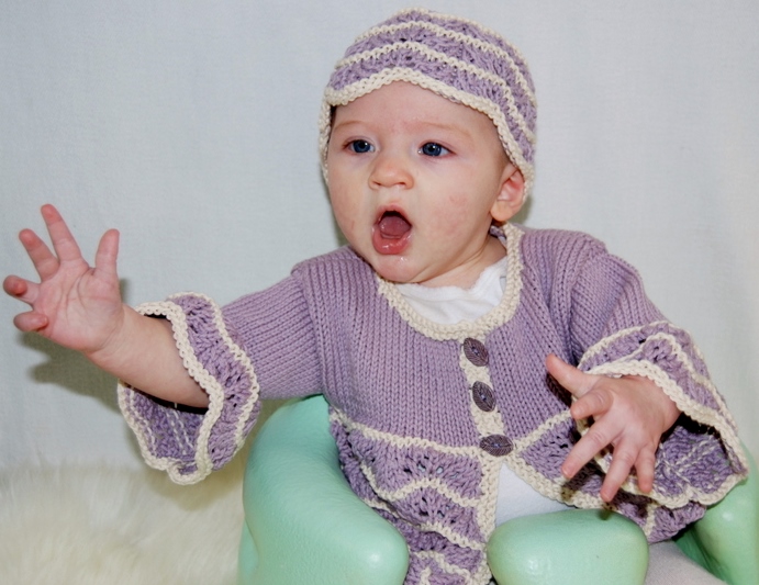A small baby sits in a pale green seat wearing a handknit lilac half-button up cardigan sweater with lace details in the hem and cuffs and a matching lacy handknit hat