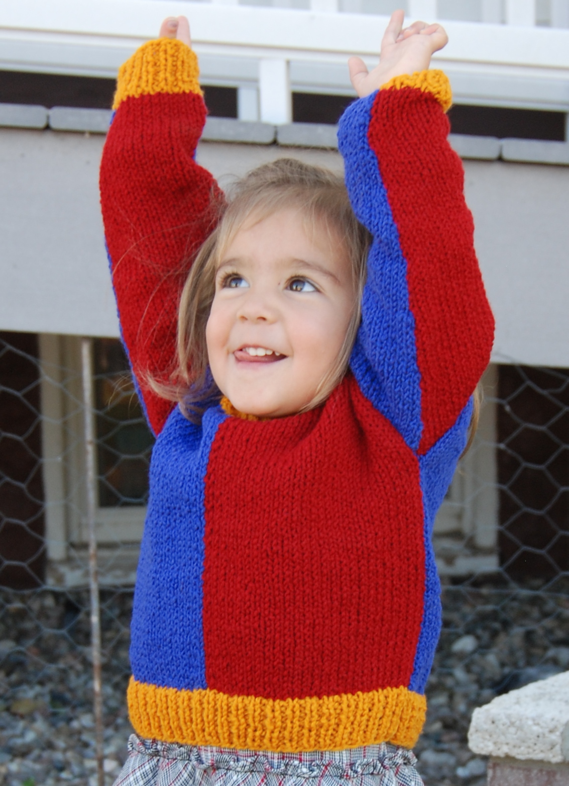 A child with raised arms wears a hand knit colorblock pullover in red, gold, and blue