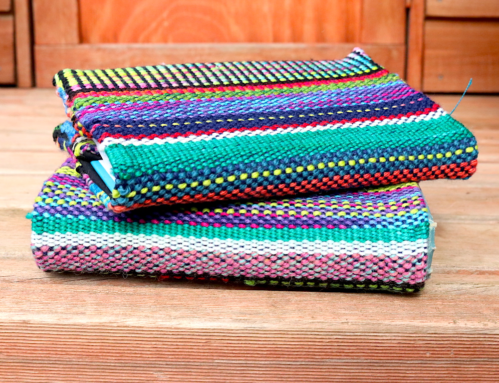 two notebooks are stacked together, each notebook has a woven cover using various colors of yarn