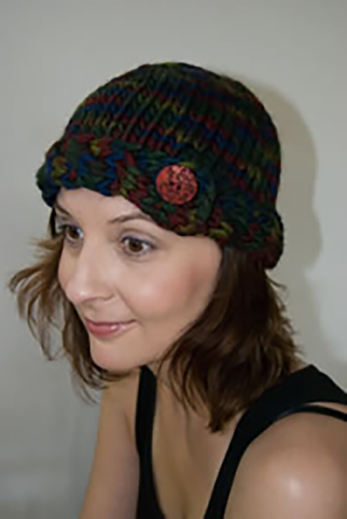 A white woman wears a dark multicolored beanie hat embellished with a large button on the brim and knit in a chunky yarn