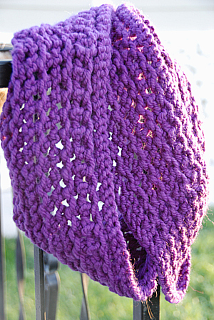 A hand knit grape purple lacy cowl hangs on a banister
