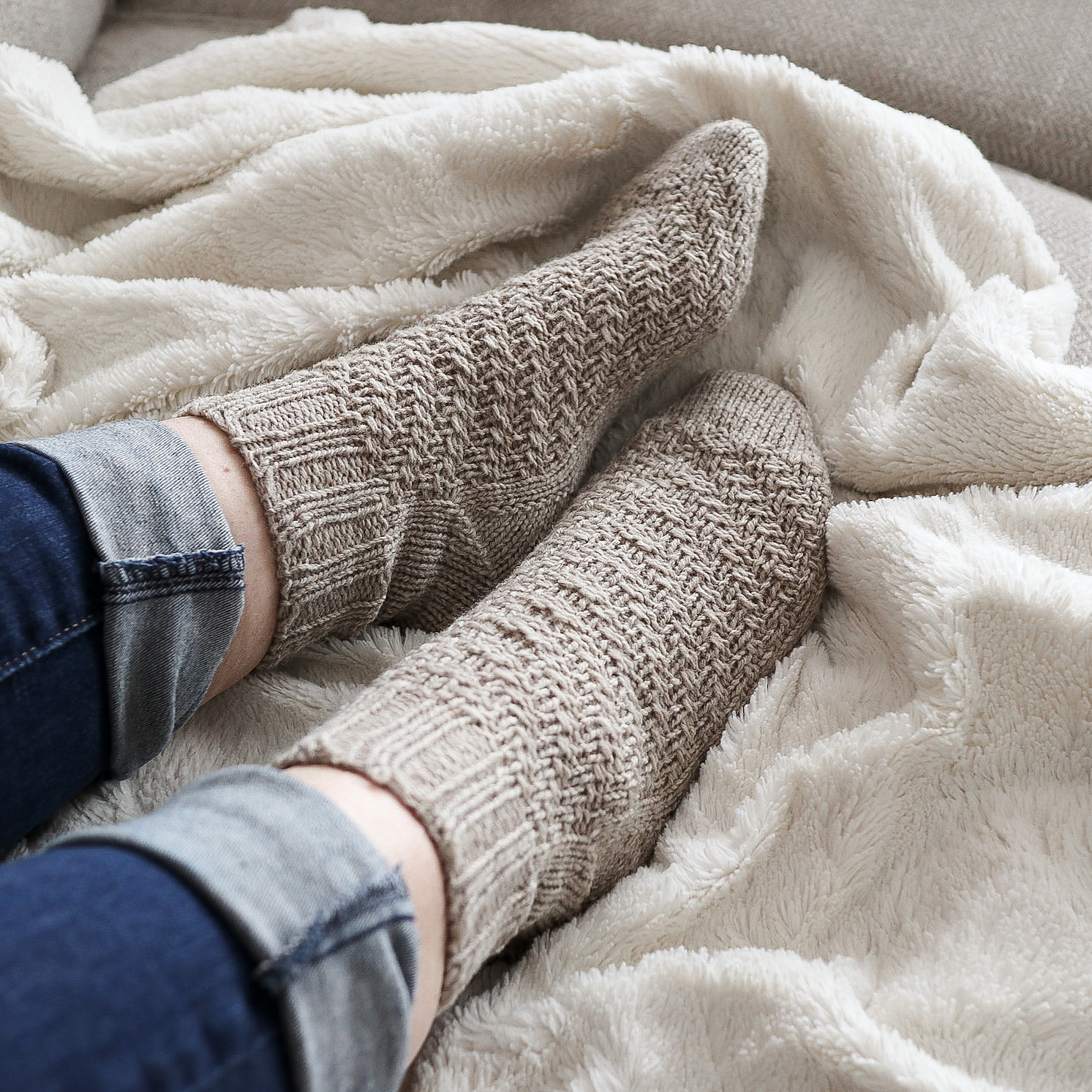 Cream colored knit socks in a wavy textural pattern worn by an unseen person rest on a white fleece blanket
