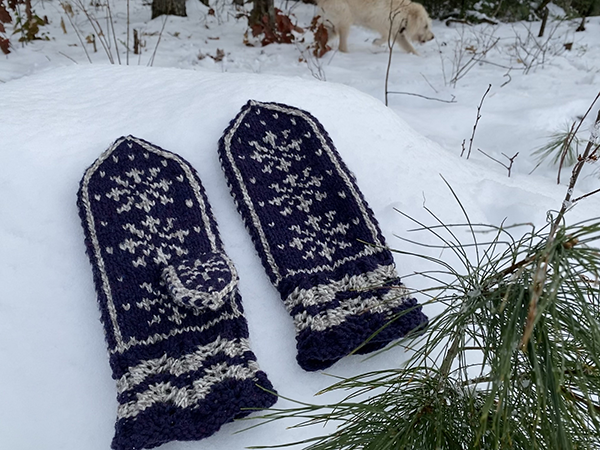 A pair of blue mittens with white colorwork snowflakes lies on the ground with fresh fallen snow