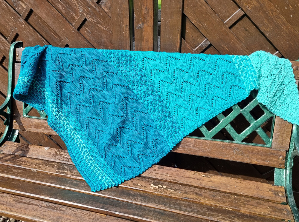 A hand knit shawl in three gradations of turquoise and featuring both colorwork and lace lies on a wooden bench