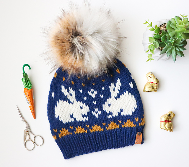 A flat lay photo of a dark navy knit hat with white colorwork bunnies and a faux fur pom pom.