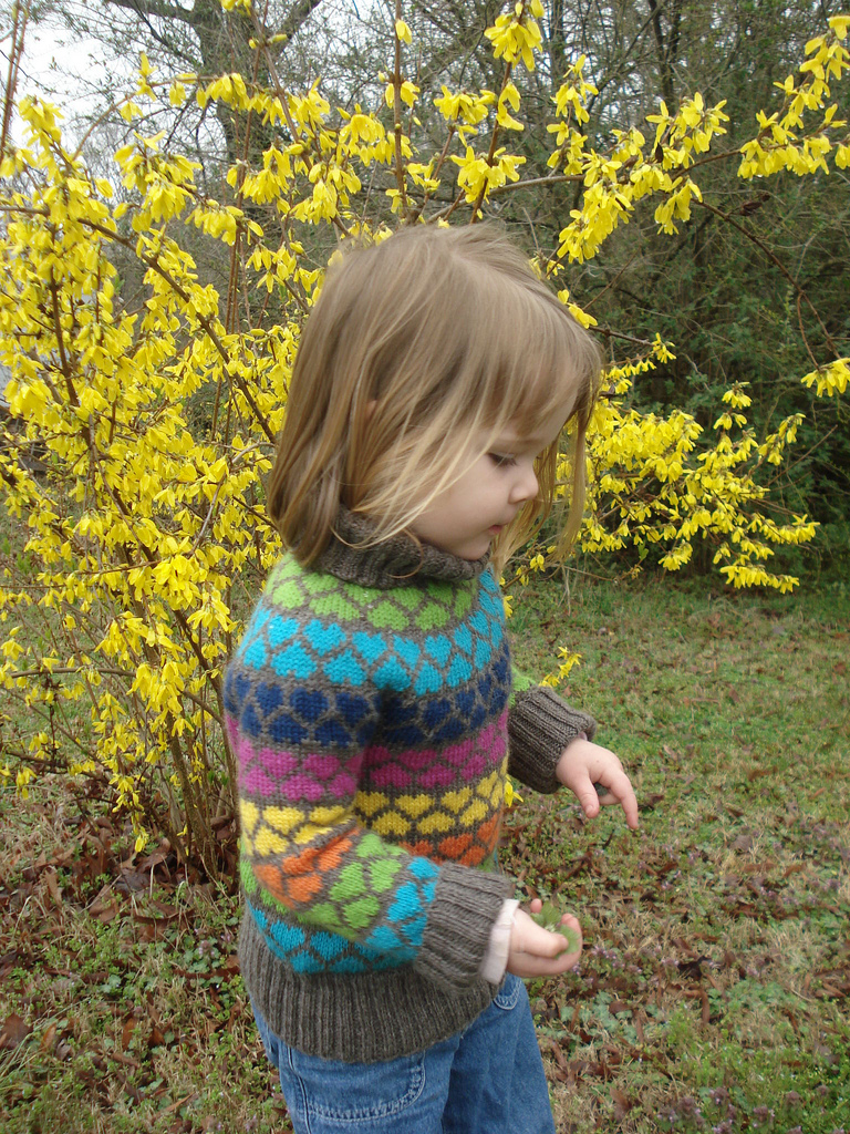 A child wears a handknit sweater featuring rows of colorwork hearts in various shades
