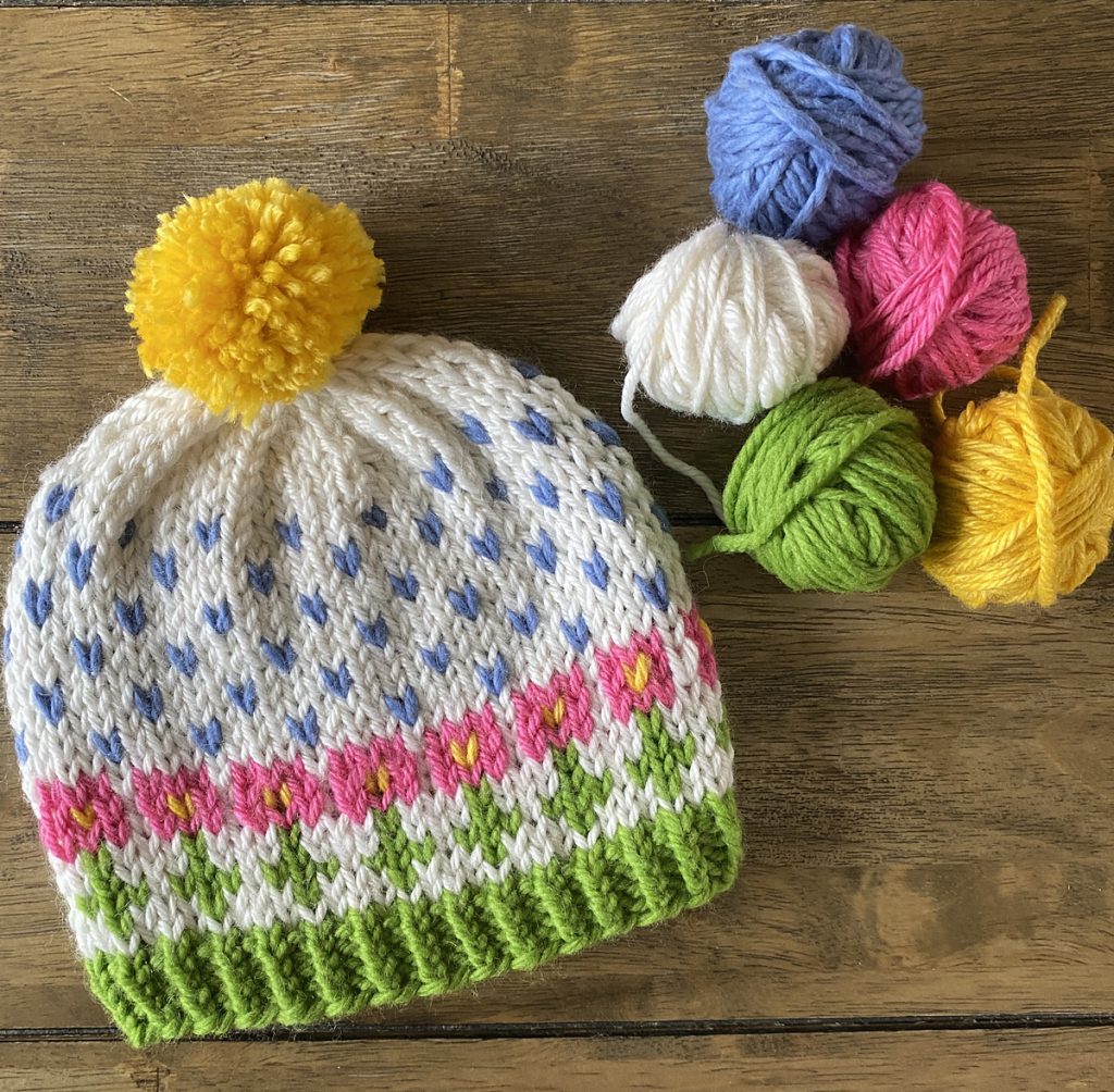 A multicolor hat with a colorwork design featuring pink flowers growing out of a green brim, blue rain falling above, topped by a yellow sun pom pom