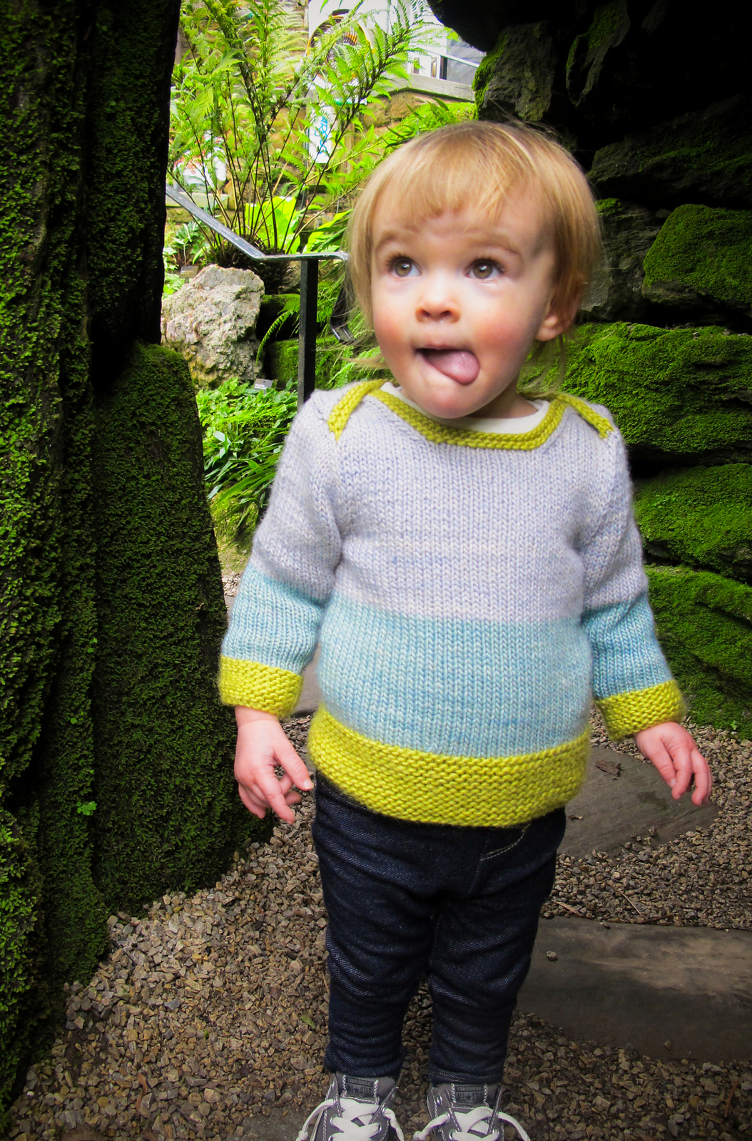A toddler wears a wide-striped hand knit sweater in pale blue, bright spring green, and french gray. The pullover features an interesting envelope-style neckline