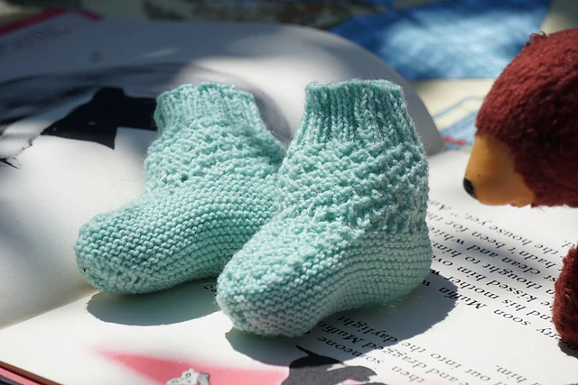 Two handknit seafoam green baby booties with moss stitch in the ankle area and a ribbed brim sit on an open book
