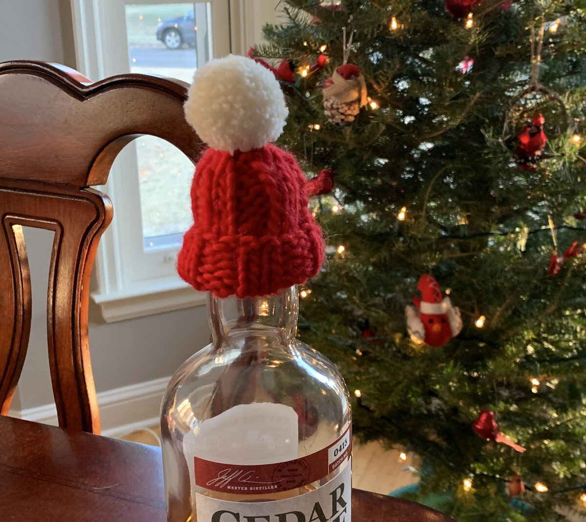 a tiny hand knit ribbed red hat with pom pom sits atop a bottle of alcohol before a holiday tree