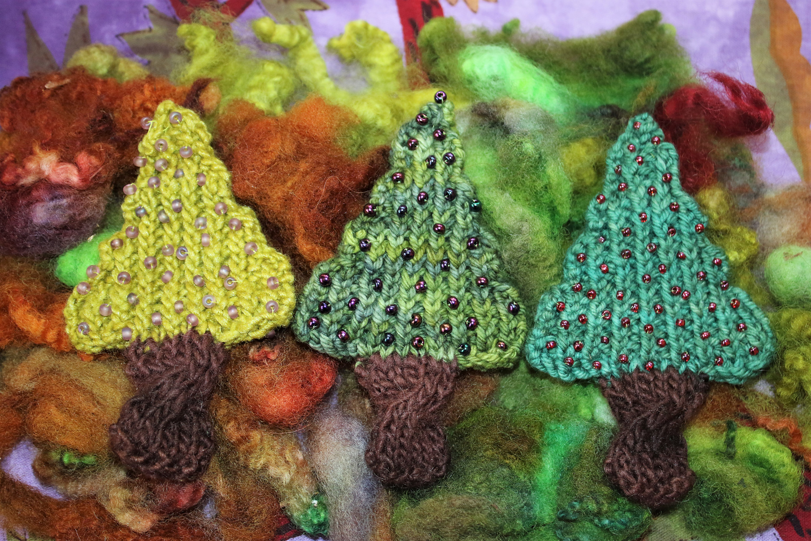 3 small handknit trees in various shades of green, decorated with beads and featuring brown cabled trunks for holiday crafting