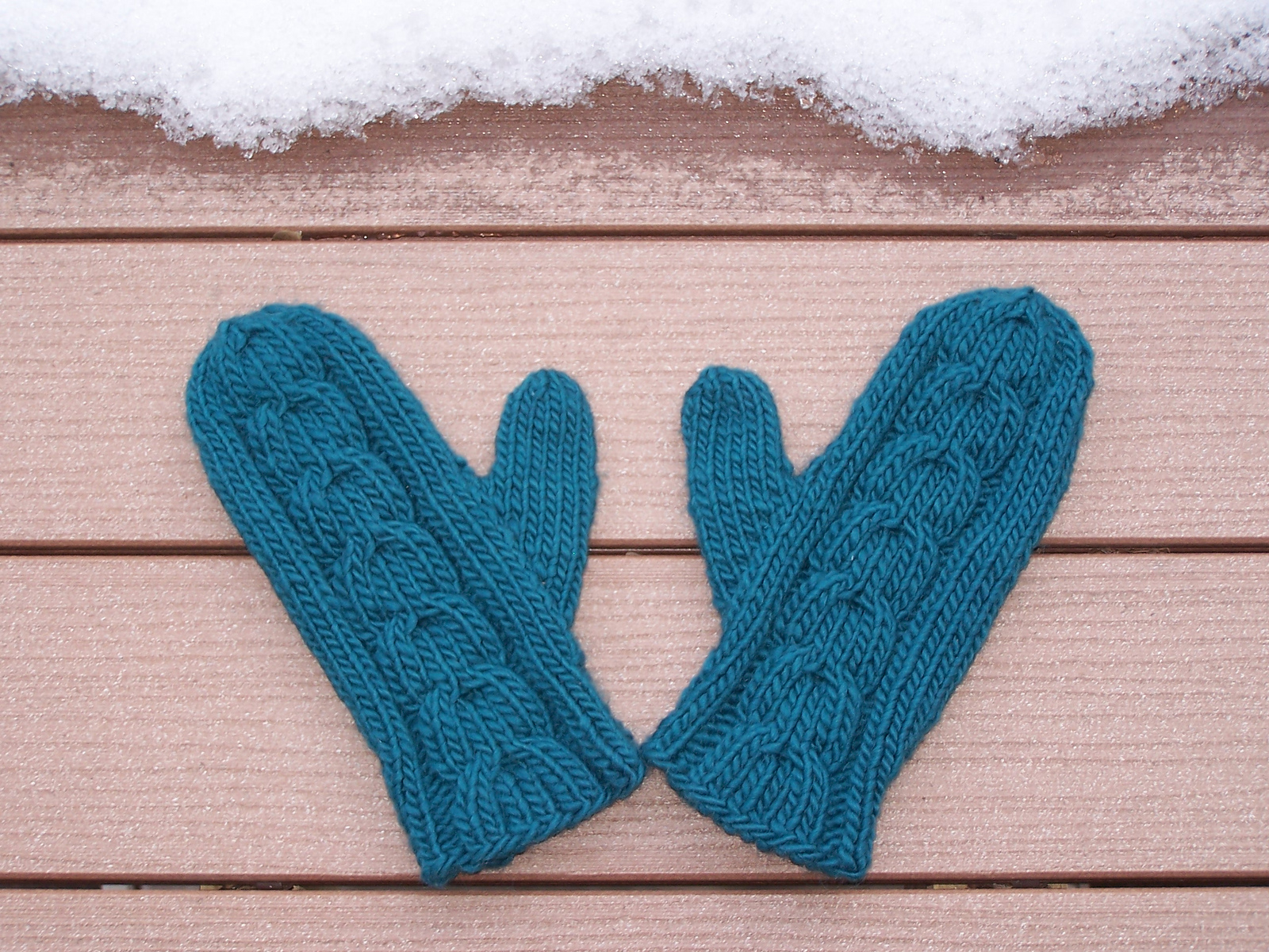 blue cabled mittens lying on a flat wood surface with winter snow