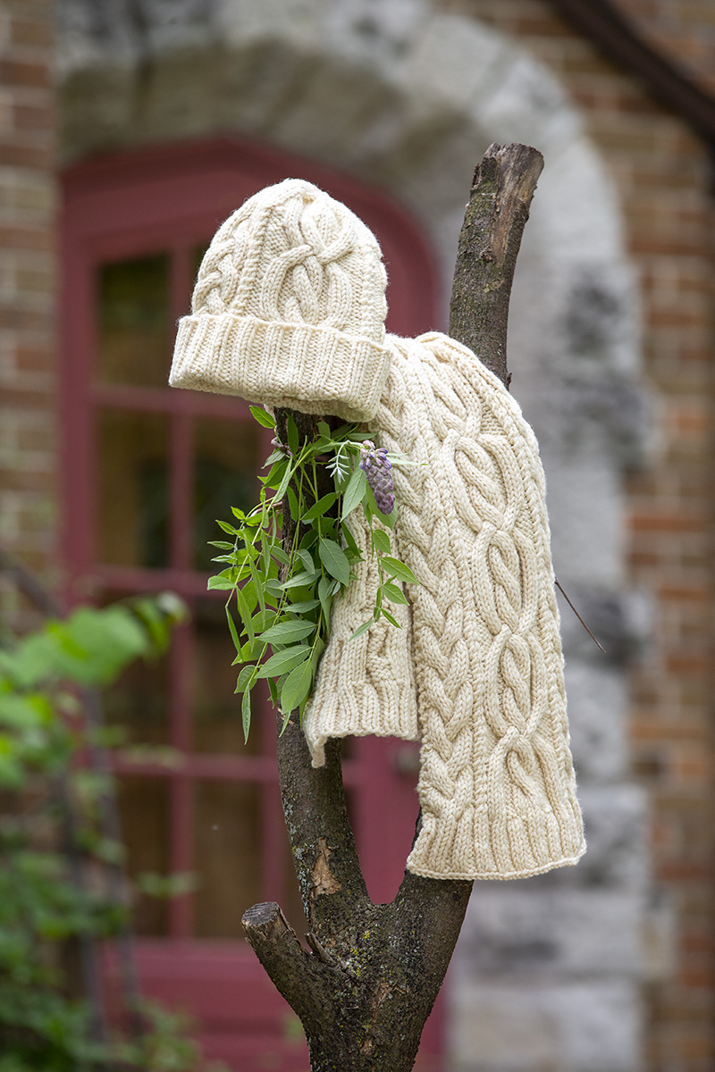 A cream colored hand knit cabled hat and scarf sit atop a leafy branch on a tree with a red door blurred in the background