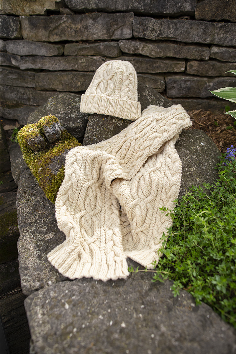 A handknit, cream colored hat and scarf with winding cables and plaits sit on a stone with moss and greenery