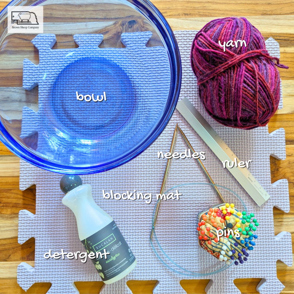 a blue bowl, blocking mat, skein of yarn, knitting needles, metal ruler, pins in a pin cushion, and small bottle of detergent