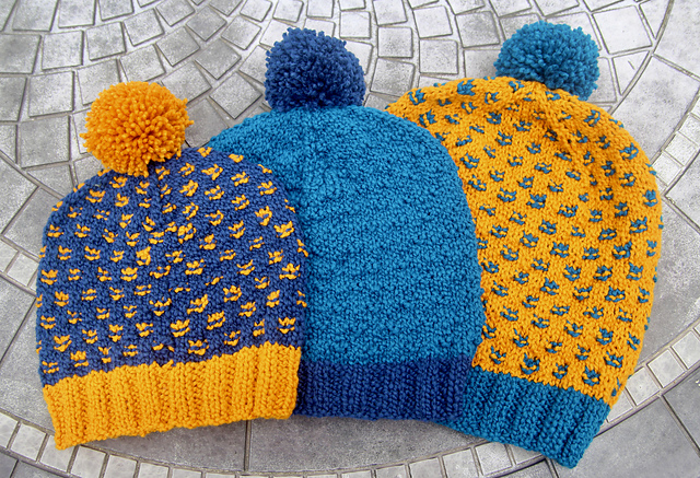 three knit colorwork hats in light blue, dark blue and yellow with contrasting pom poms