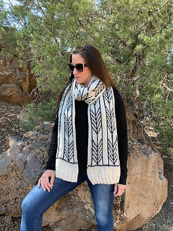 A woman and a black shirt and sunglasses wears a hand knit white scarf with a black arrow head design