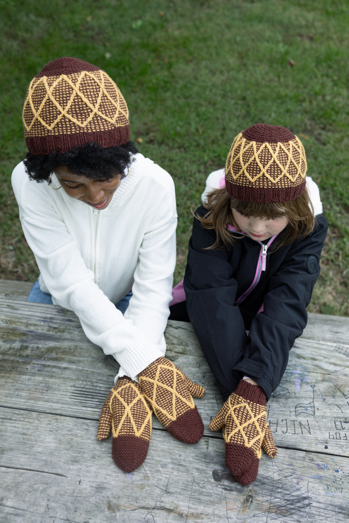 A bird's eye view of a young woman and a young girl wearing matching burgundy and yellow colorwork hats and mittens in a twisted stitch trellis design